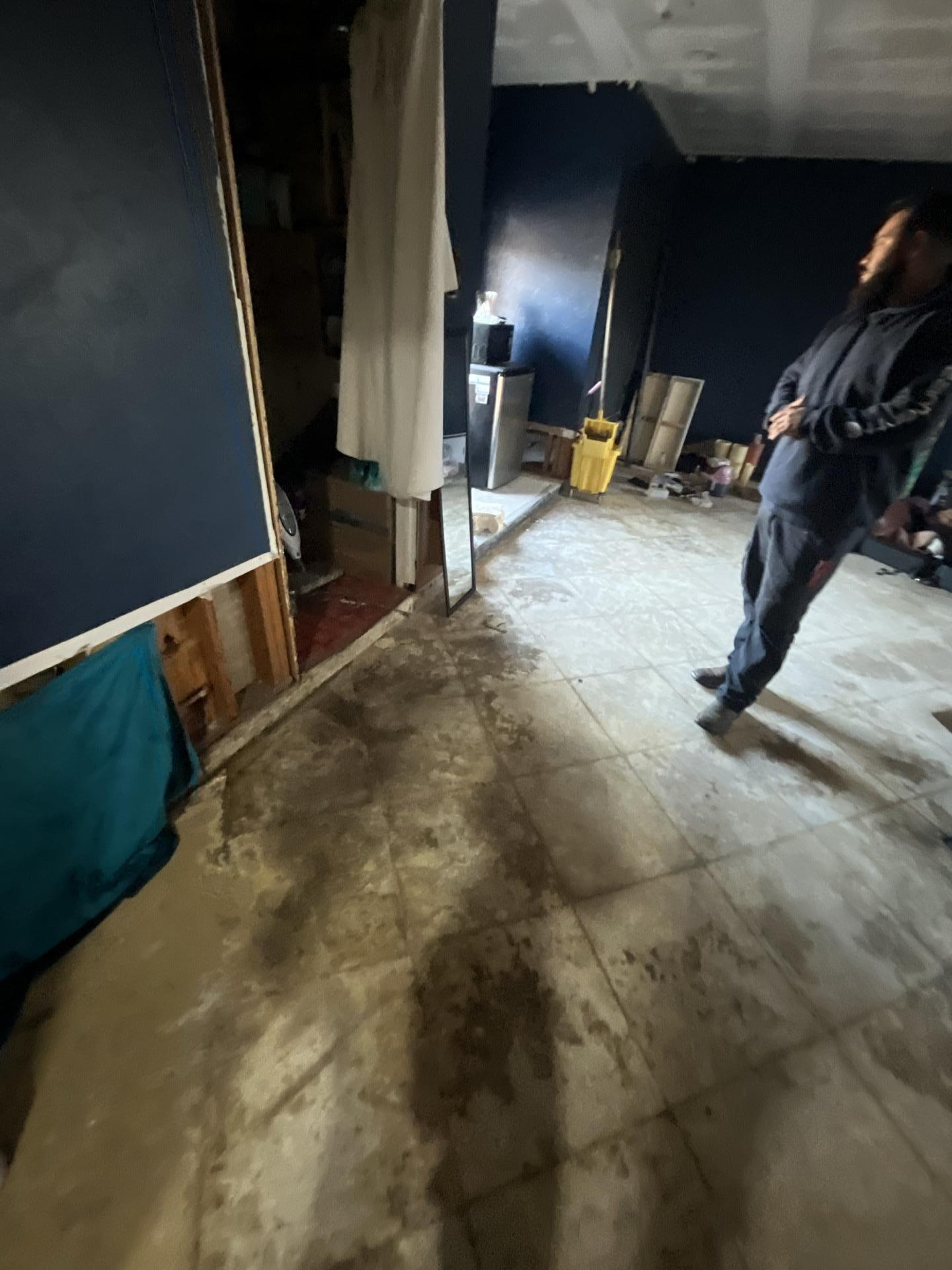 Wide view of a living room with significant water damage on floor tiles and exposed walls, a technician assessing the restoration progress.
